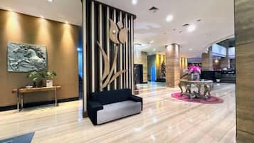 Un vestíbulo de hotel con un sofá negro, una mesa redonda con camino de mesa rosa y un panel decorativo en la pared.