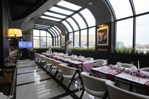 Un restaurante con techo curvo y ventanas amplias, mesas dispuestas con manteles blancos y sillas, y una pantalla de televisión.