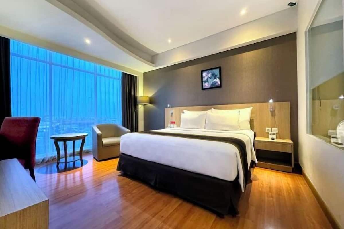 A modern hotel room with a large bed, a desk, a chair, and a view of the city through large windows.