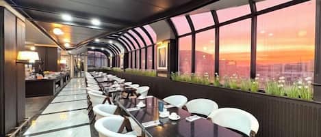 Une grande salle à manger avec des tables et des chaises, de grandes fenêtres offrant une vue sur la ville et un aménagement intérieur moderne.