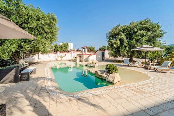 Una piscina con cubierta de baldosas, rodeada de árboles y algunas sillas de descanso.