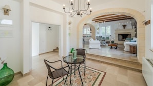 Un amplio salón con una mesa de comedor, sillas y una araña. Un arco de entrada lleva a la cocina y a la chimenea.