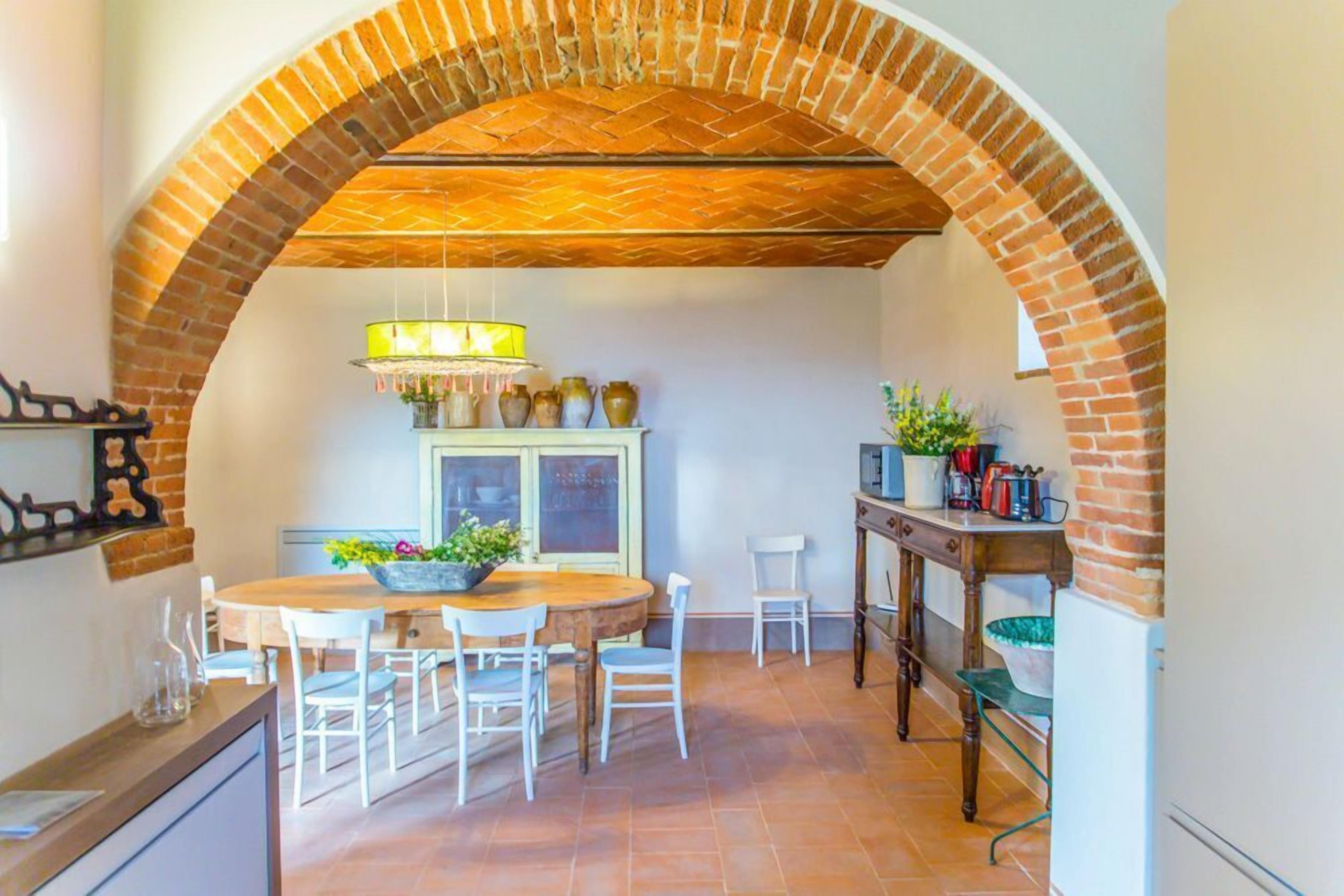 A dining area with a brick archway, a wooden table, white chairs, and a hanging light fixture.