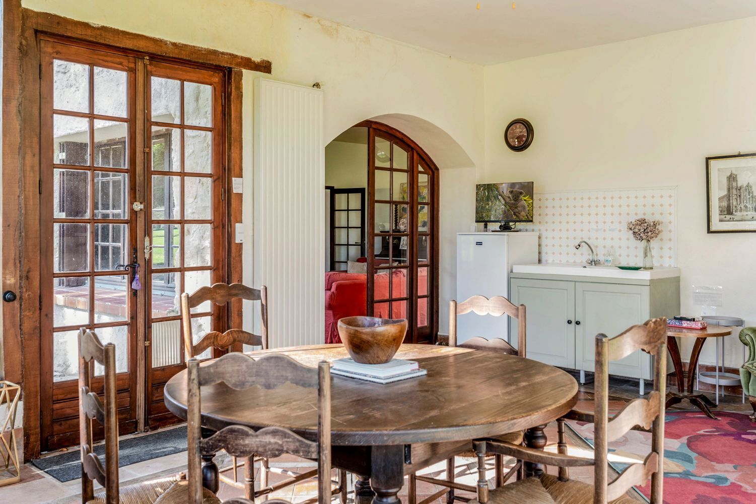 A dining area with a round wooden table, chairs, a large bowl, and a view of a red sofa through an arched doorway.