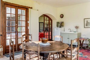 A dining area with a round wooden table, chairs, a large bowl, and a view of a red sofa through an arched doorway.