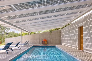 Una piscina al aire libre con zona techada, sillas de descanso y un muro de concreto.