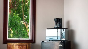 Une cuisine compacte avec une machine à café posée sur un réfrigérateur, une poubelle et une fenêtre donnant sur un espace verdoyant.