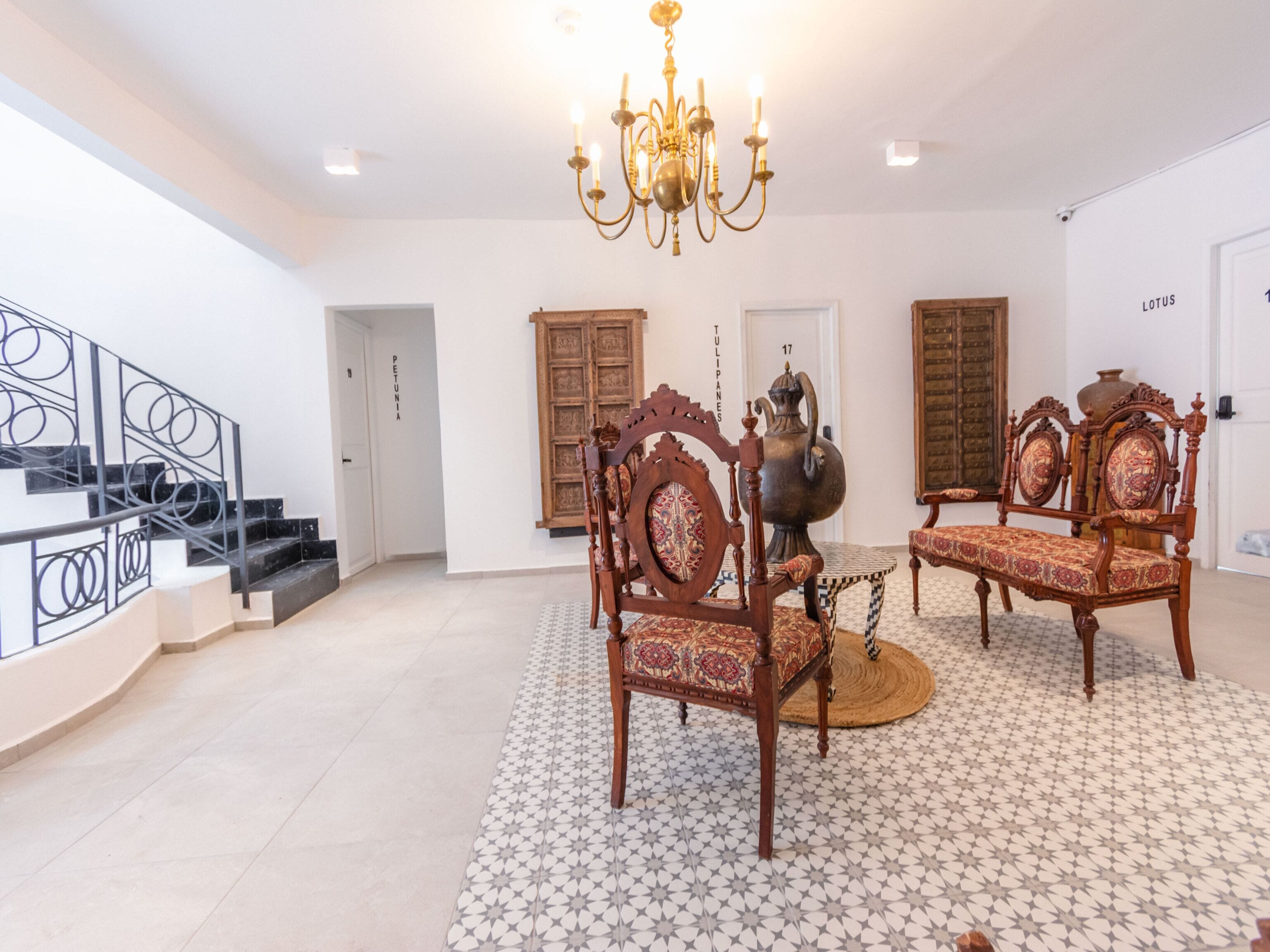 A spacious room with a chandelier, two ornate wooden chairs, a patterned rug, and a staircase with black railings.