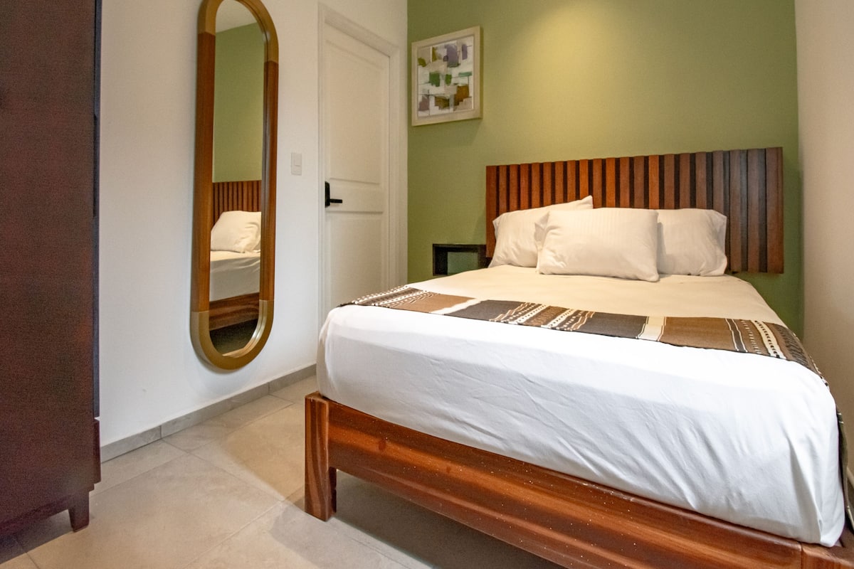 A neatly made bed with white linens and a patterned throw, a wooden headboard, a large mirror, and a door with a white frame.