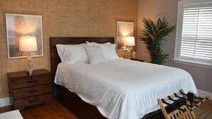 Un dormitorio ordenado con una cama grande, dos mesitas de noche con lámparas, una silla de madera y una planta en maceta.