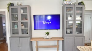 Una sala de estar con un televisor montado en la pared, armarios grises con puertas de cristal y una mesa de madera en el centro.