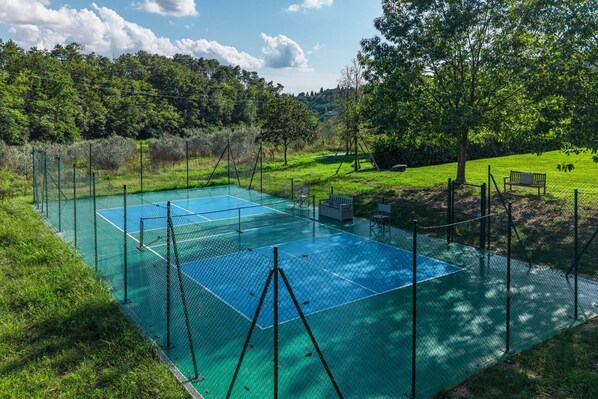 Una cancha de tenis al aire libre rodeada por una cerca, con bancos y árboles de fondo.