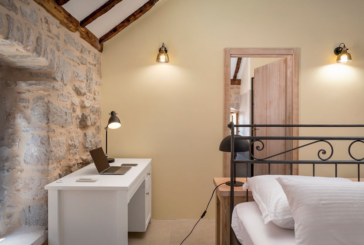 A bedroom with a stone wall, a wooden desk with a laptop, a bed with white linens, and two wall-mounted lights.