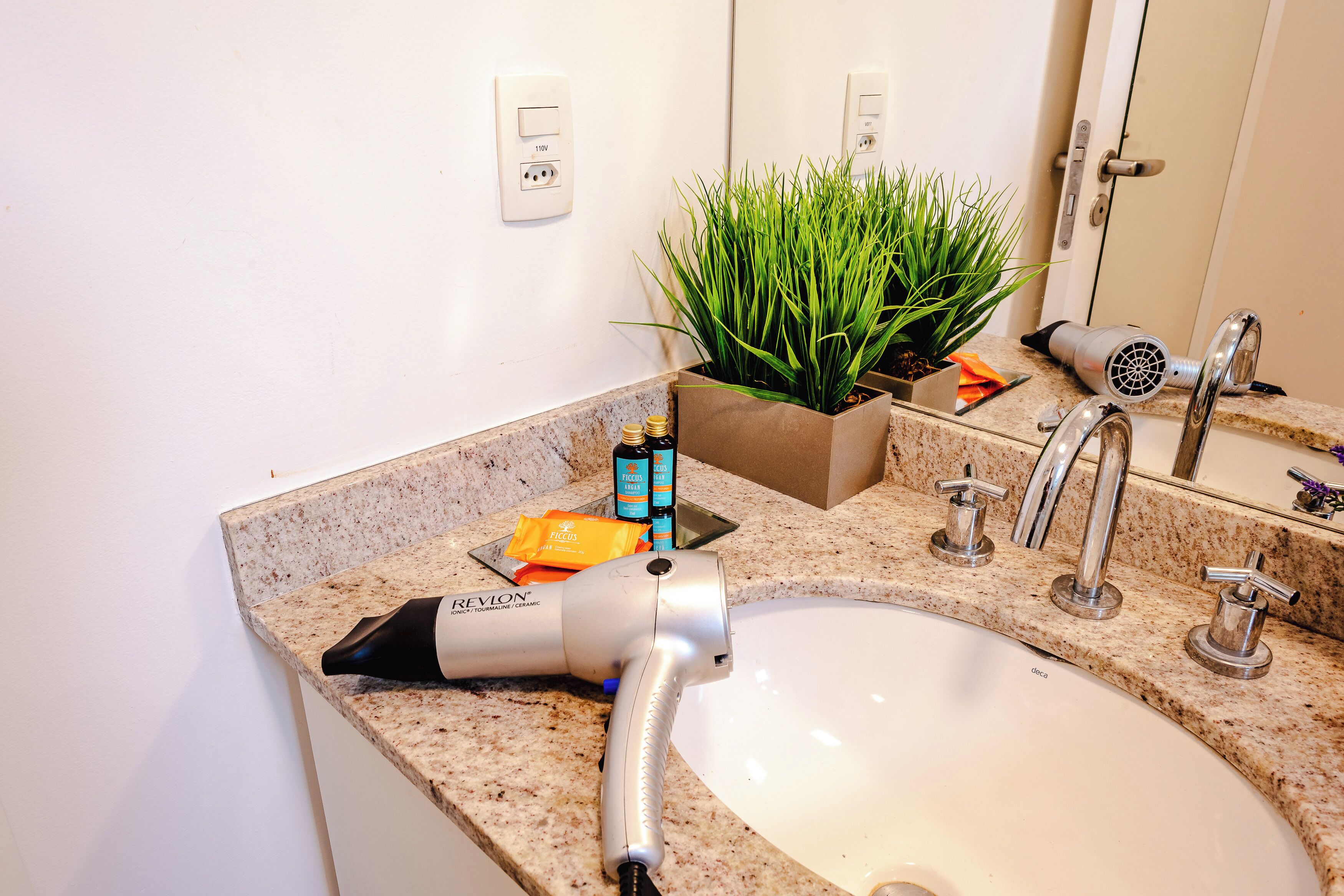 A bathroom with a sink, a hairdryer, a plant, and toiletries on the countertop.