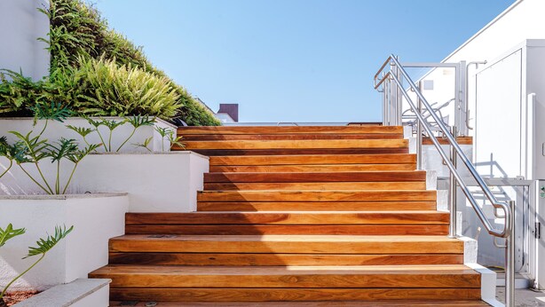 Una escalera con peldaños de madera y barandillas metálicas que conduce a un edificio blanco con jardín en la azotea.