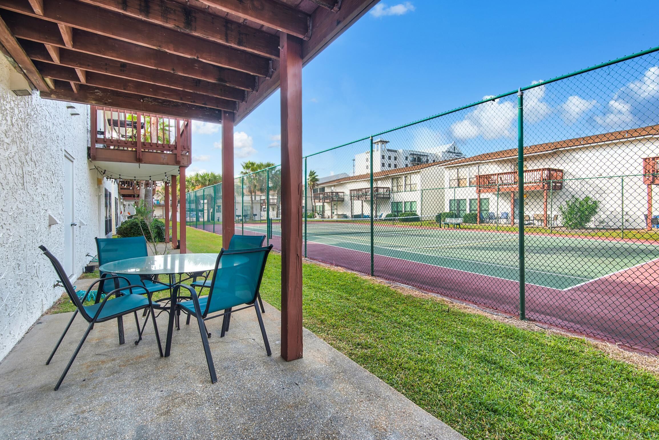 A covered patio with a table and chairs, a tennis court, and a building with balconies in the background.