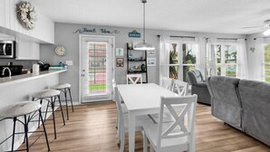 A modern kitchen with white cabinets, a dining area with a white table and chairs, and a grey sofa in a bright living room. - Beachview 107 (Navarre)