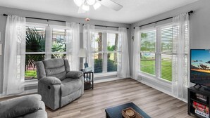 A spacious living room with a ceiling fan, large windows with blinds, a grey sofa, a small table, and a TV on a stand. - Beachview 107 (Navarre)