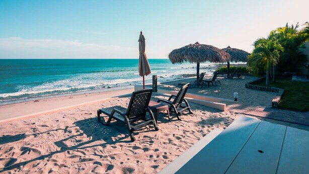 Eine Strandkulisse mit Liegestühlen, einem Tisch, einem Reetdachschirm und Blick auf das Meer.