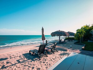 Eine Strandkulisse mit Liegestühlen, einem Tisch, einem Reetdachschirm und Blick auf das Meer.
