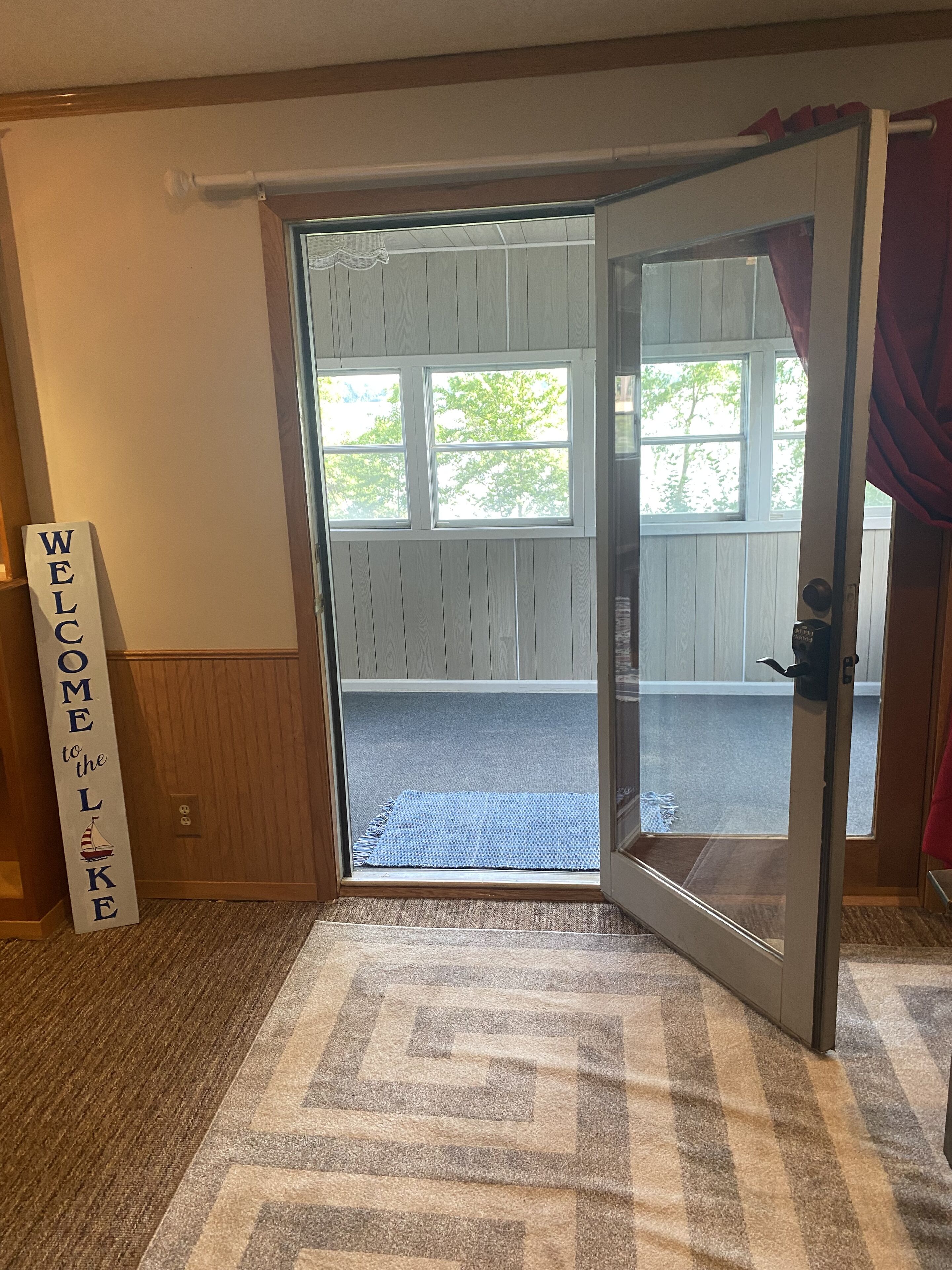 A room with a sliding glass door leading to an outdoor area with a view of trees. There is a welcome sign on the wall and a rug on the floor.