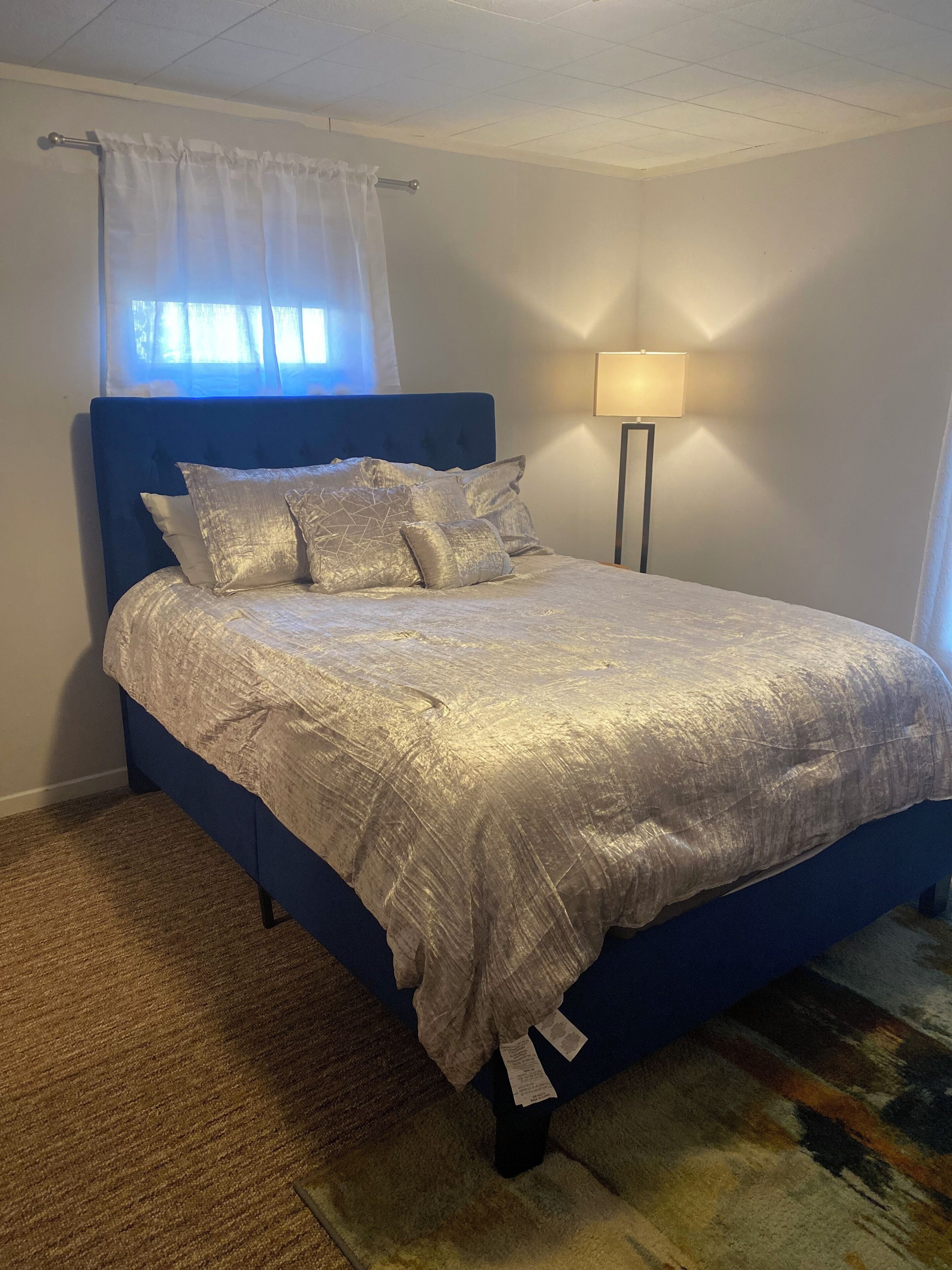 Ein Schlafzimmer mit einem blauen Kopfteil, einem Bett mit silberner Decke und Kissen, einer Stehlampe und einem Fenster mit weißem Vorhang.