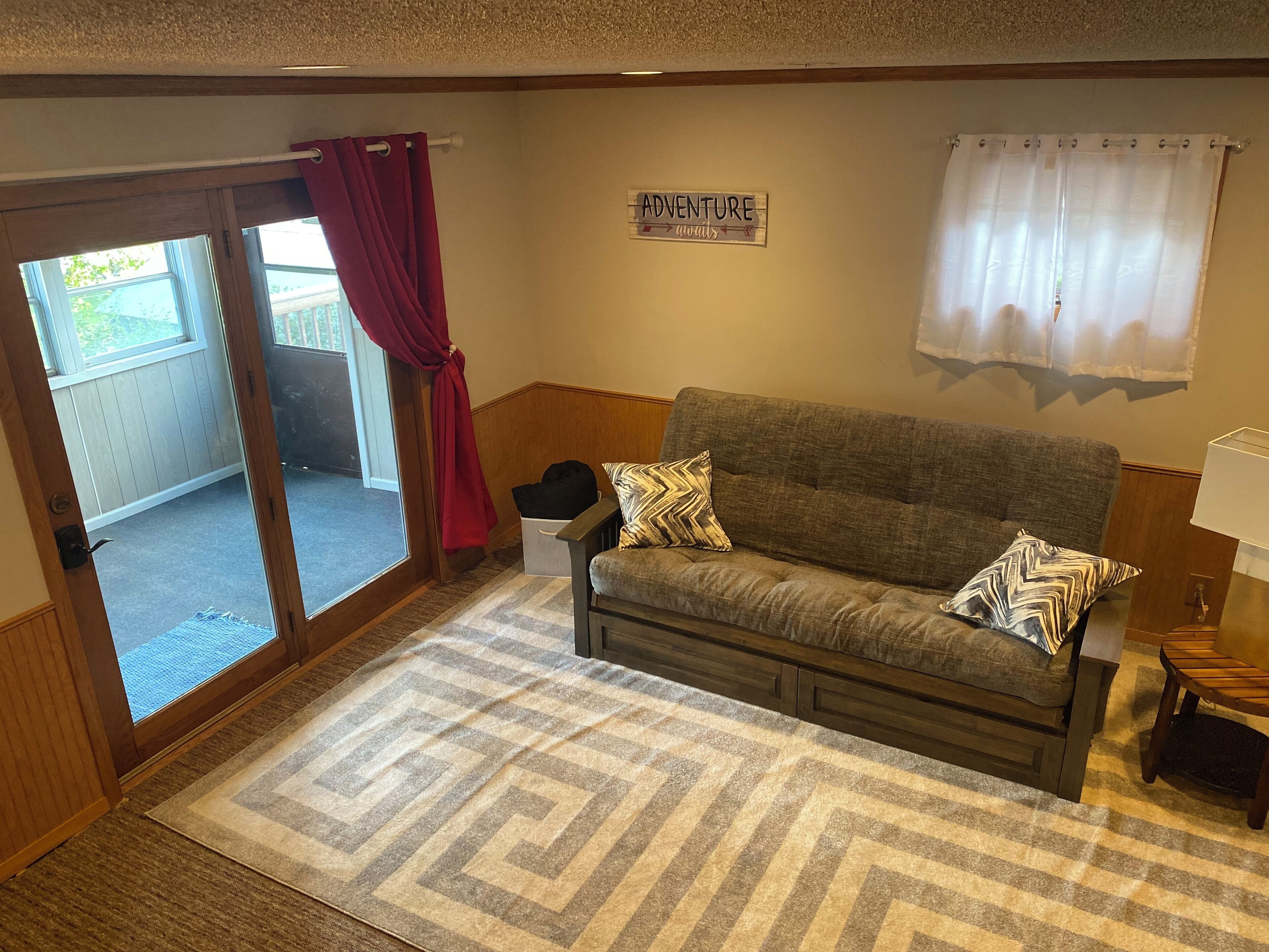 A cozy living room with a grey sofa, patterned throw pillows, a wooden coffee table, and a rug. A sign on the wall reads "ADVENTURE awaits."