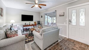 A living room with two grey sofas, a ceiling fan, a flat-screen TV, a coffee table, and a window with a view of trees.