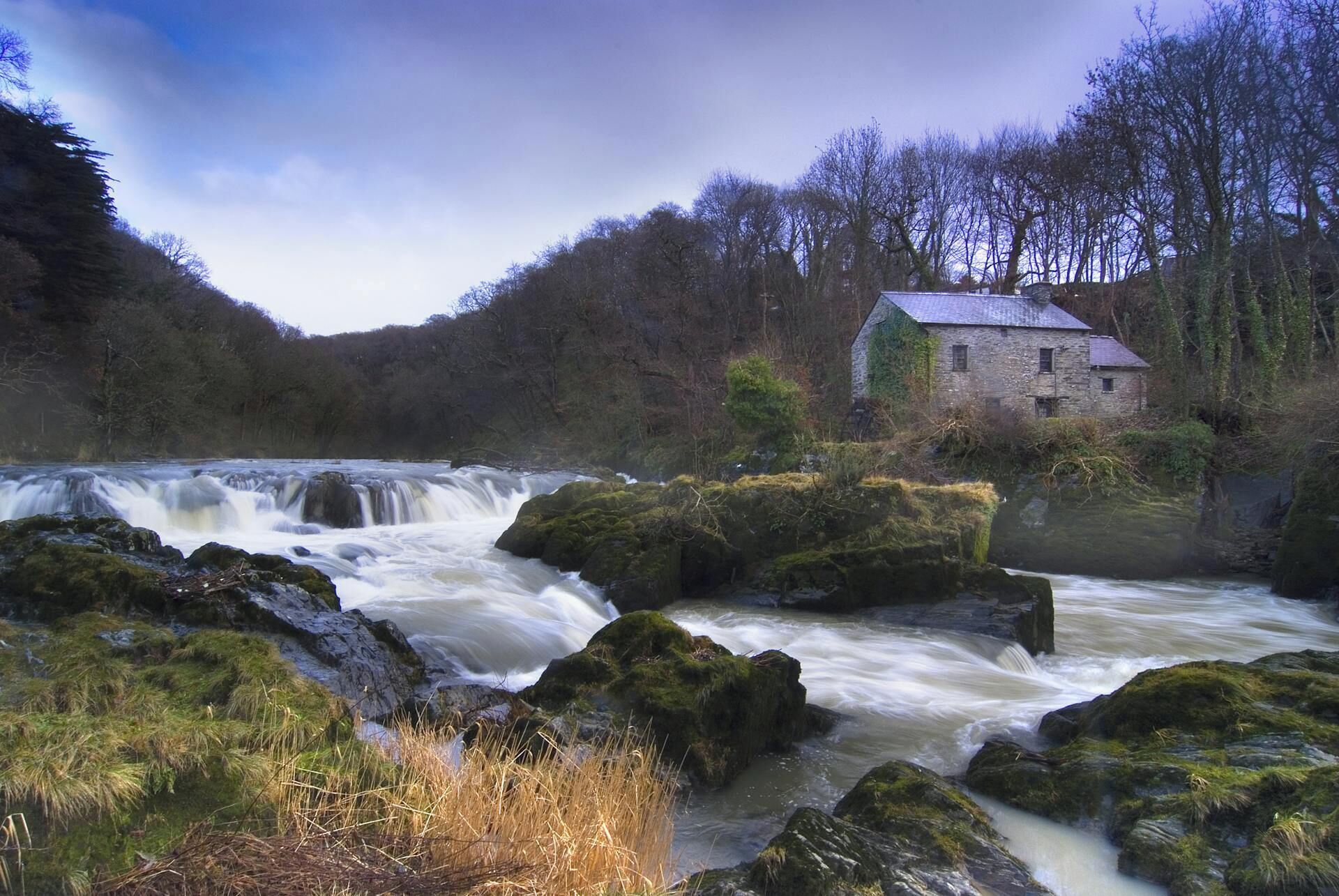 A river with a waterfall, surrounded by rocks and vegetation, and a stone building with a sloped roof.
