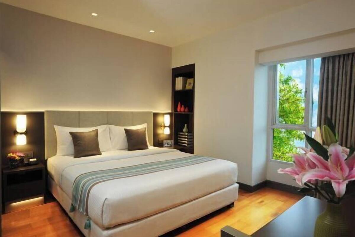 Une chambre d’hôtel moderne, dotée d’un grand lit, de lampes de chevet, d’une table de chevet, d’une bibliothèque et d’une fenêtre donnant sur un paysage verdoyant.