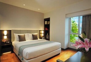 Una habitación de hotel moderna con una cama grande, lámparas de noche, una mesita de noche, una estantería y un ventanal con vistas a zonas verdes.