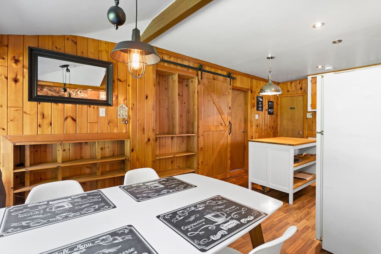 Un intérieur rustique en bois, avec un espace repas comprenant des tables blanches et des sets de table noirs et blancs, un grand miroir et des étagères en bois.