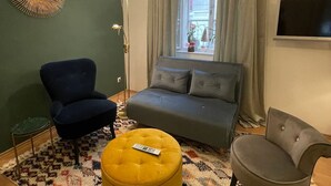 Ein Wohnzimmer mit einer Couch, zwei Sesseln, einem gelben Hocker, einer Stehlampe und einem an der Wand befestigten Fernseher.