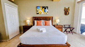 A neatly made bed with a white duvet, two wooden nightstands with lamps, a colorful painting above the bed, and a wooden chair with a table beside the window. - Conch – Oceanfront Ground-Floor Studios – Panama (San Carlos)