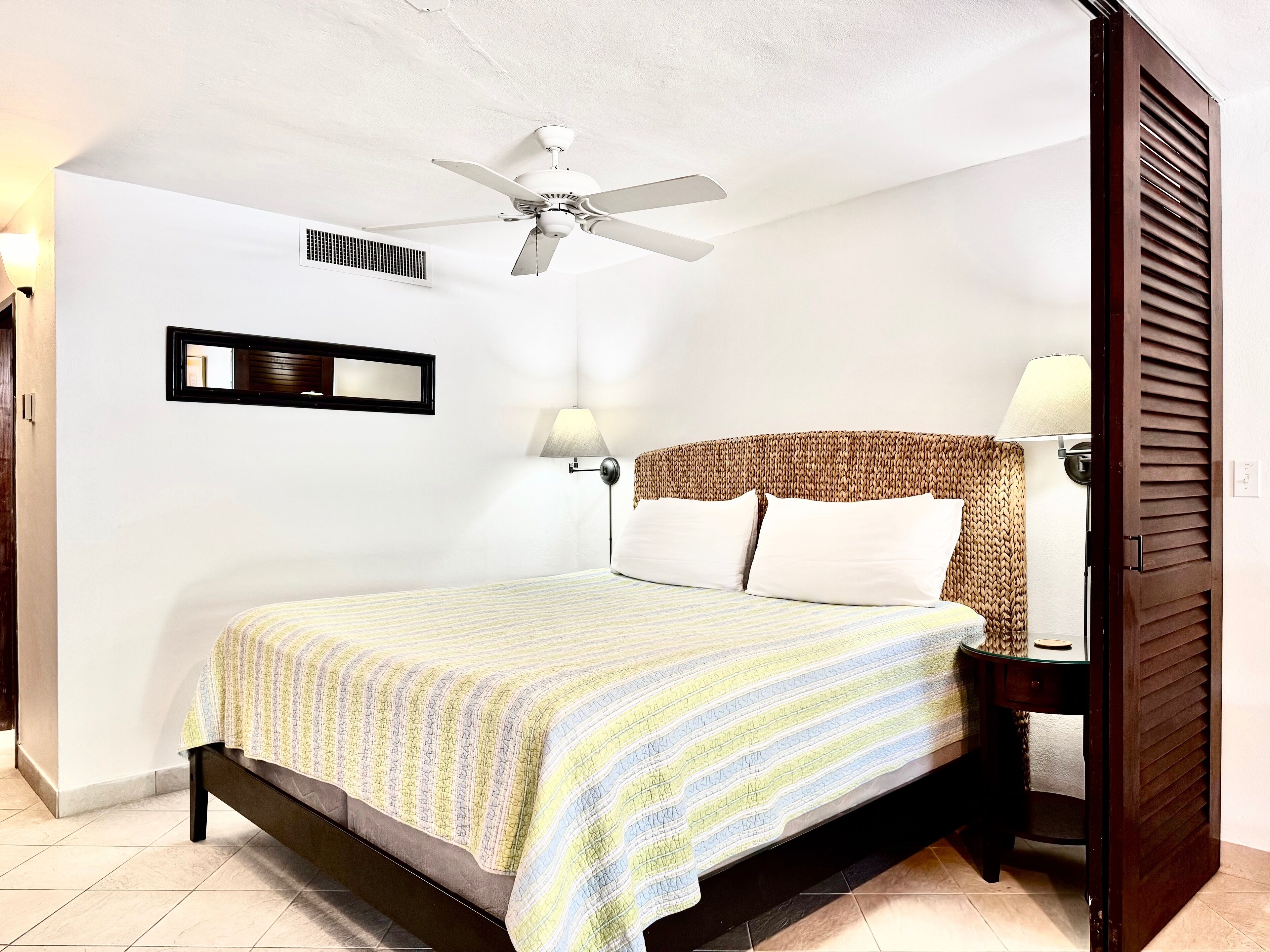 Un dormitorio ordenado con una cama, un ventilador de techo, una mesita de noche con lámpara y un cabecero de madera.