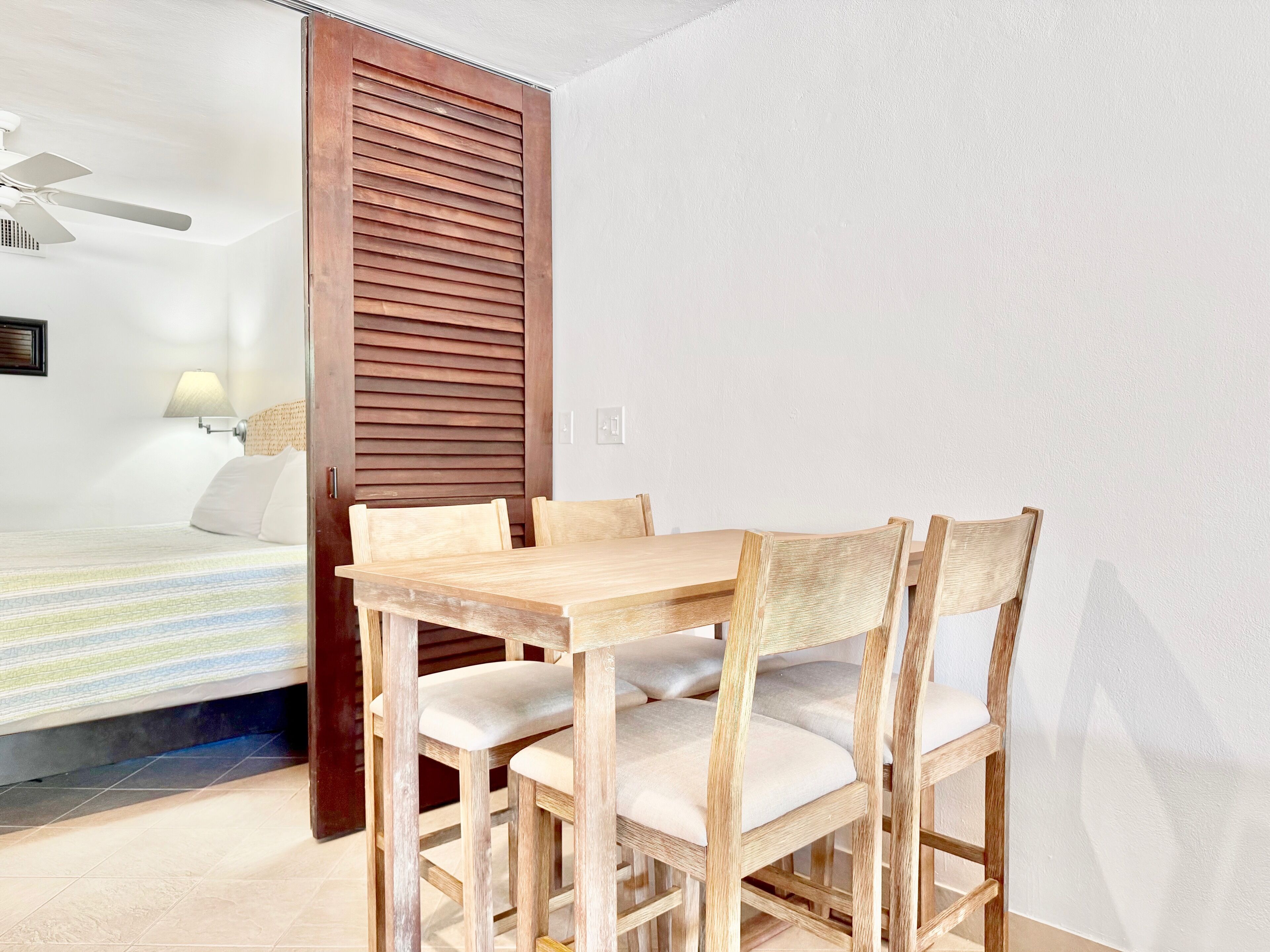A hotel room with a wooden dining table and chairs, a bed with a striped bedspread, a ceiling fan, and a wall-mounted lamp.