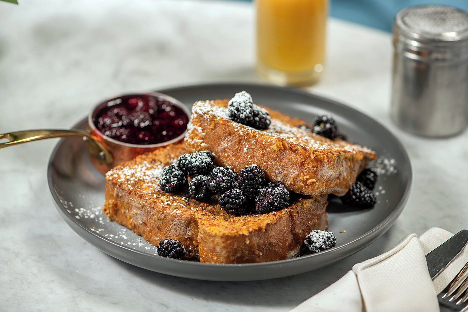 A plate of French toast with blackberries and powdered sugar, accompanied by a bowl of berry compote.