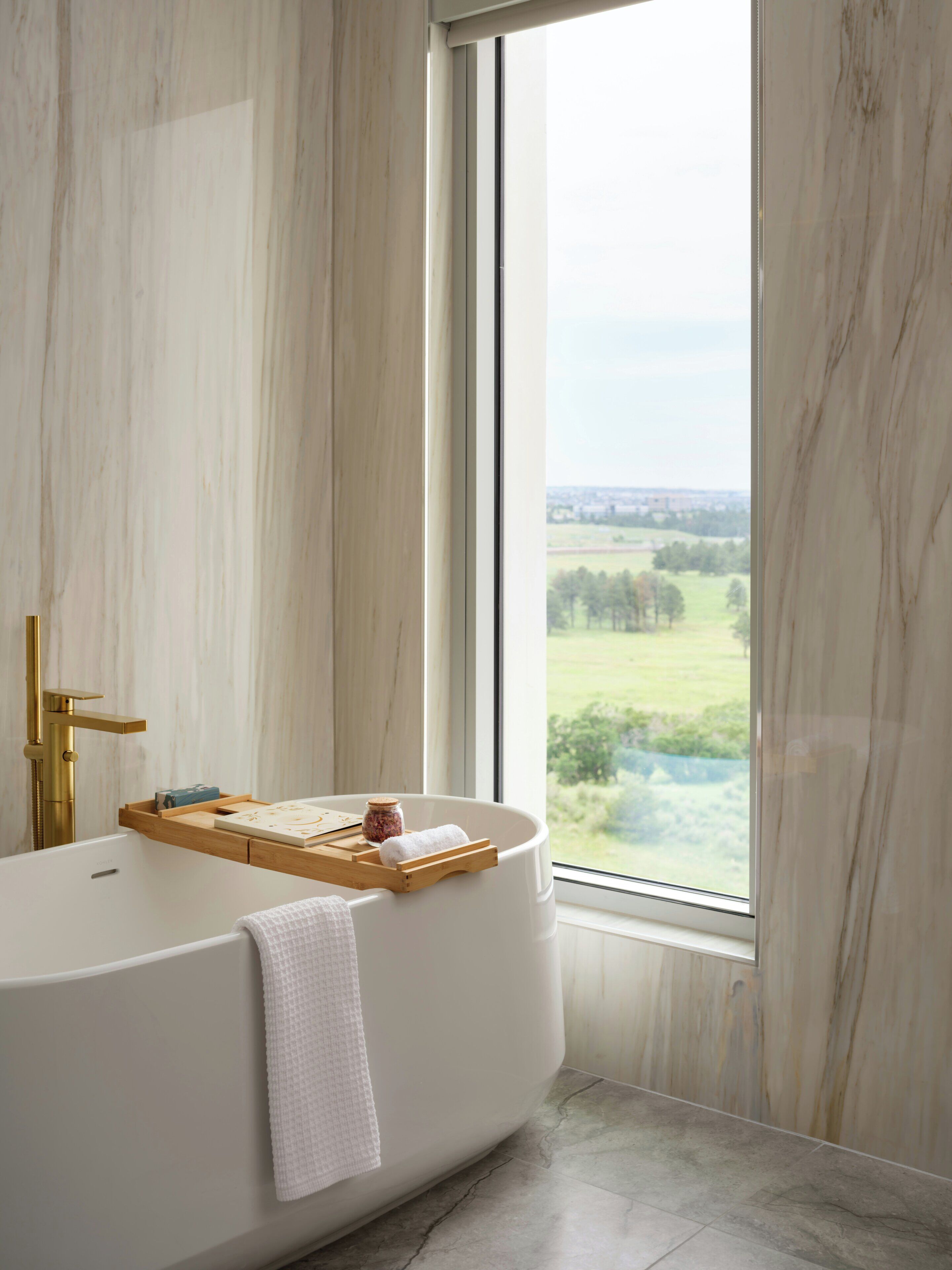 A modern bathroom with a freestanding bathtub, a gold faucet, a wooden tray, and a view of a green landscape through a large window.