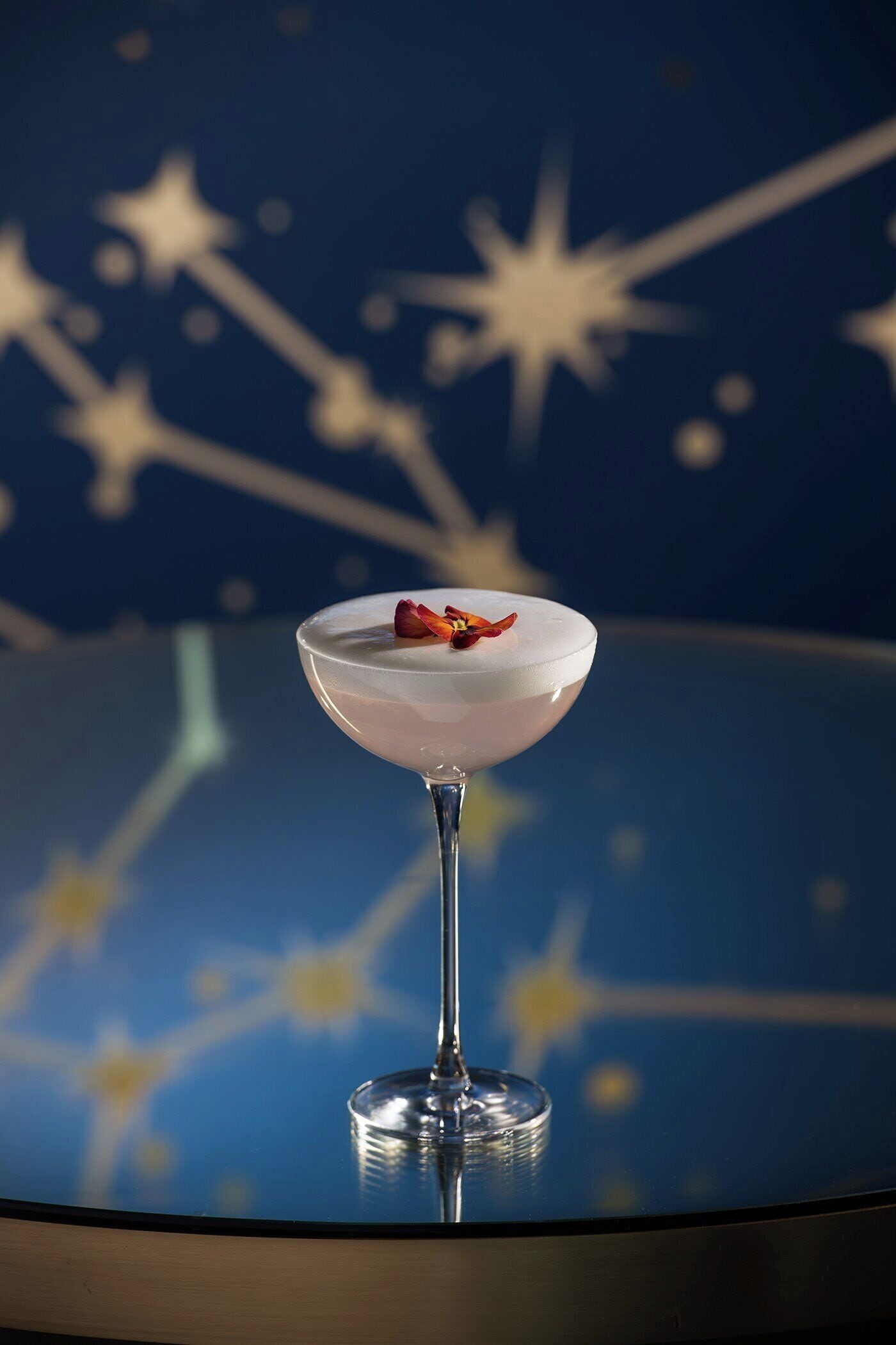 A martini glass with a pink drink and a flower on a reflective surface with a starry background.