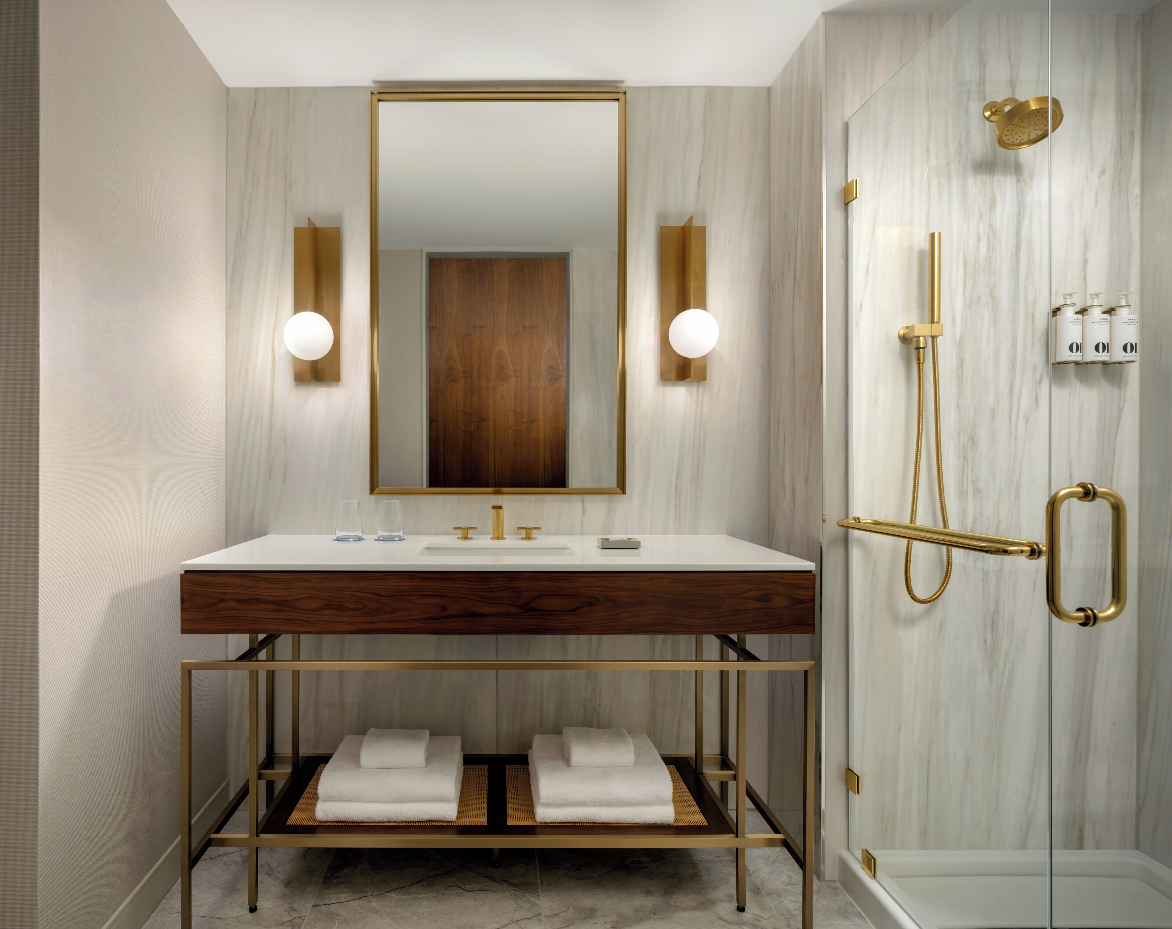 Une salle de bain moderne avec un meuble-lavabo en marbre, un grand miroir, une pomme de douche dorée et une douche à parois de verre.