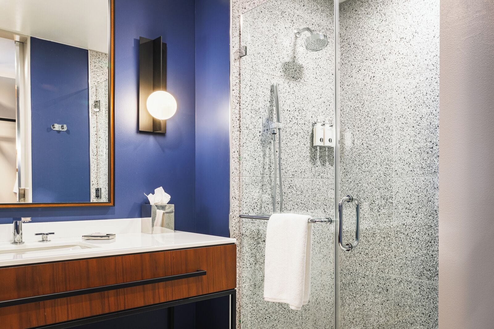Une salle de bain avec un mur bleu, un meuble-lavabo en bois, un miroir rond, un porte-serviettes et une douche dont le mur est recouvert de carreaux à motifs.