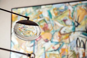 Una lámpara colgante moderna con pantalla de cristal transparente y base de metal, suspendida sobre un cuadro abstracto colorido.