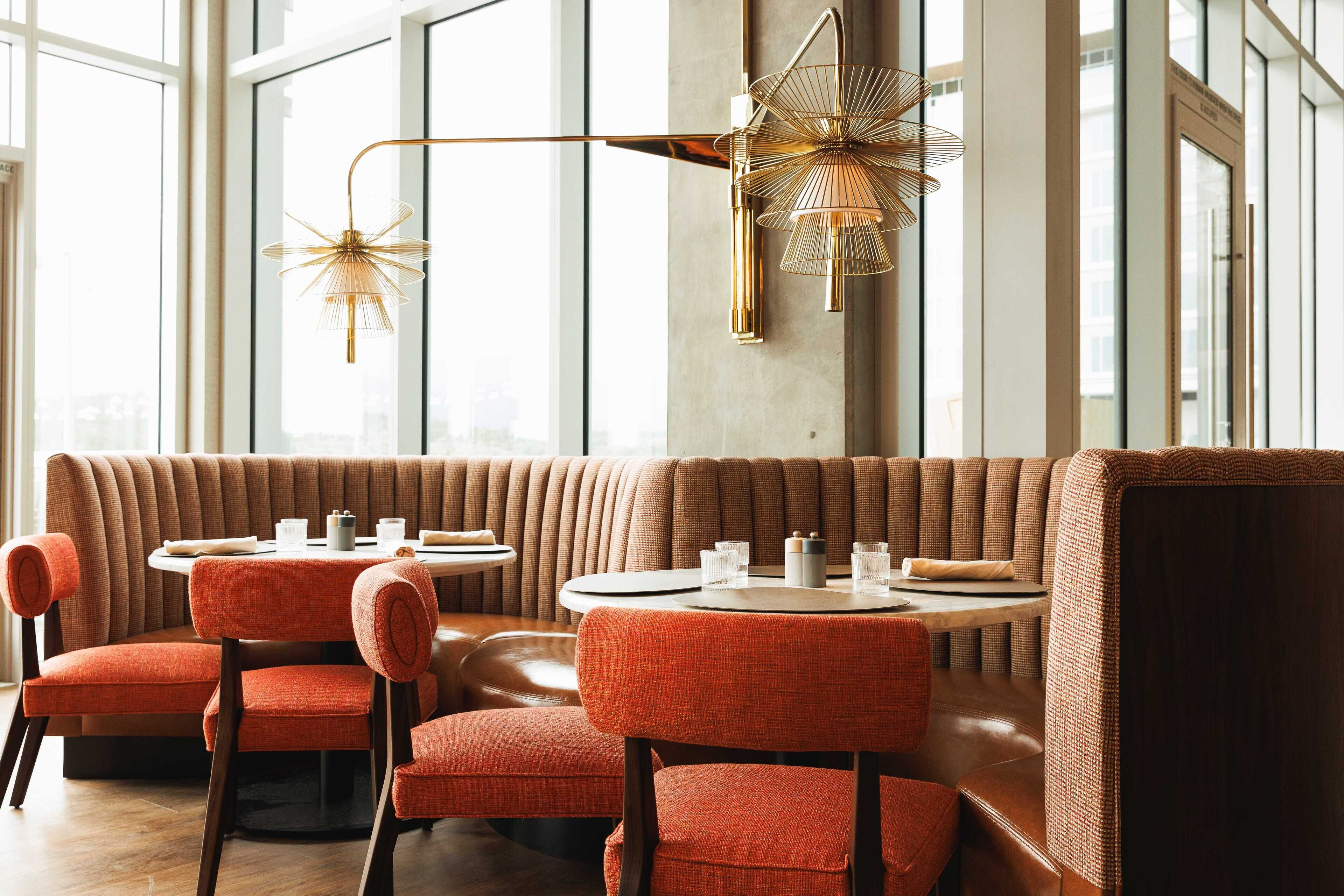 Un restaurante moderno con techo alto, ventanas amplias y un área de asientos con mesas redondas y sillas acolchadas.