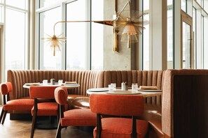 Ein modernes Restaurant mit hoher Decke, großen Fenstern und einer Sitzgelegenheit mit runden Tischen und gepolsterten Stühlen.
