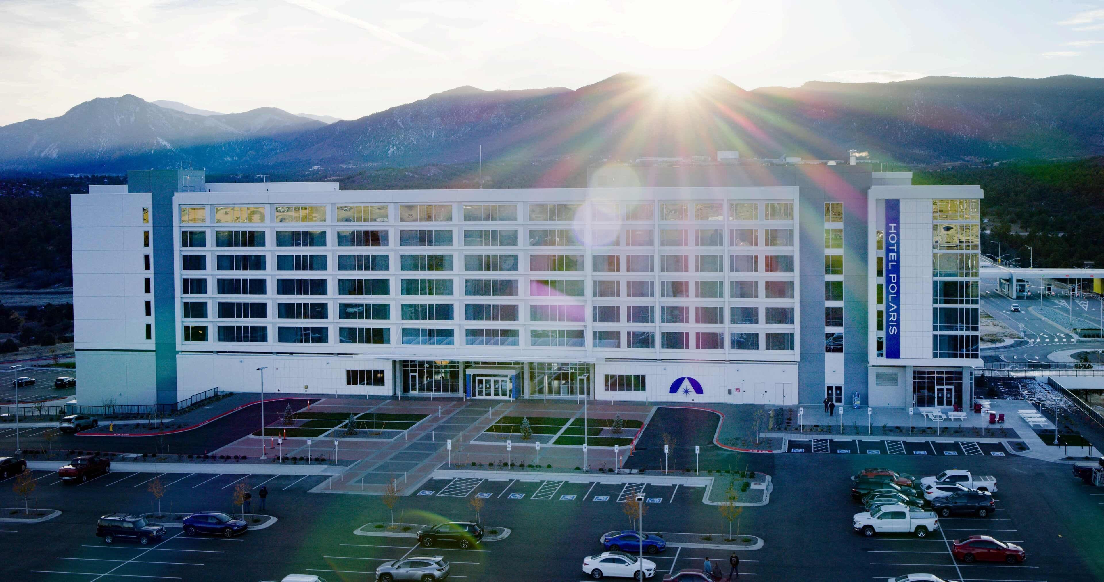 Ein modernes Hotel mit einem zentralen Eingang, mehreren Parkplätzen und Blick auf Berge im Hintergrund.