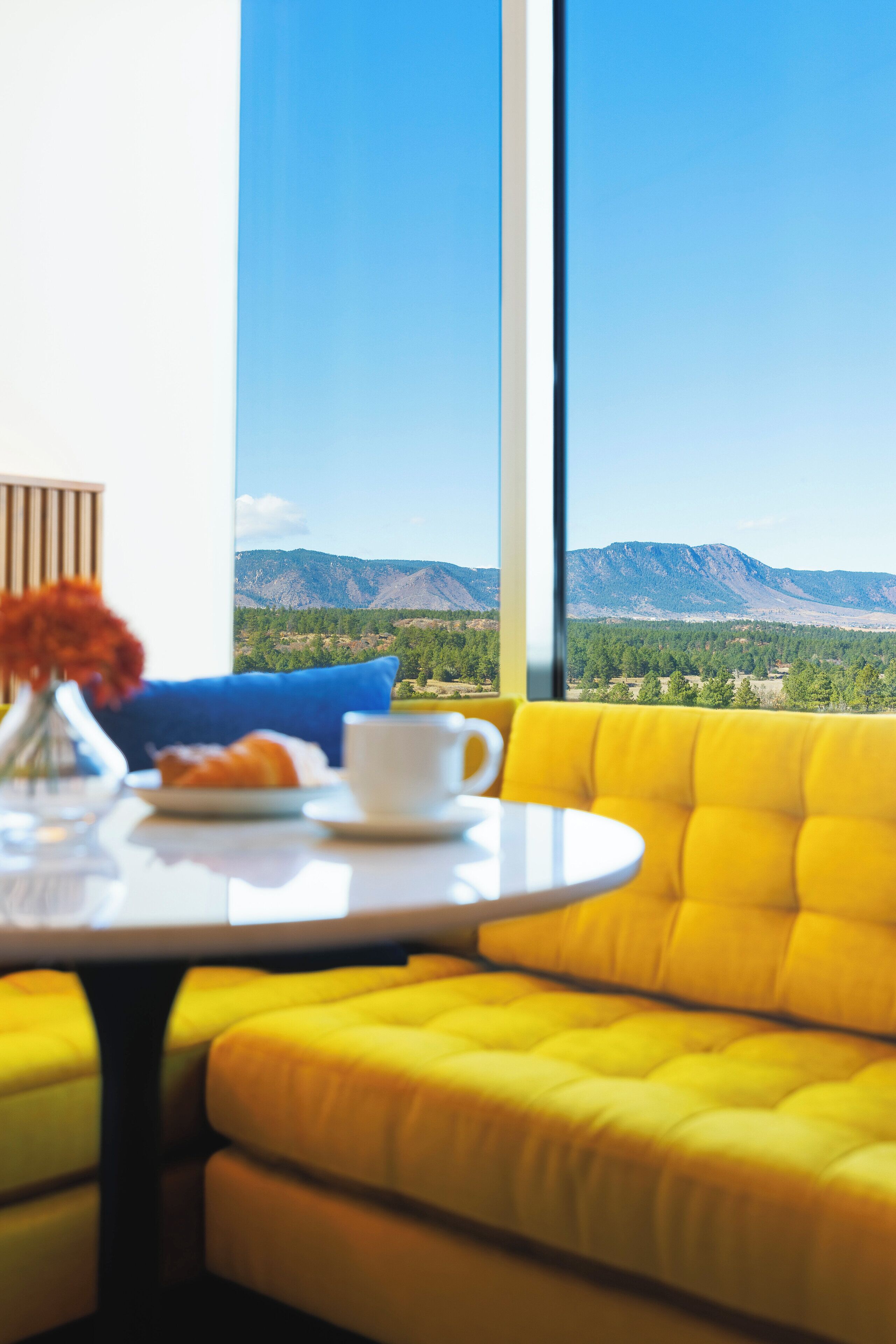 A room with a yellow sofa, a small round table, and a view of mountains through large windows.