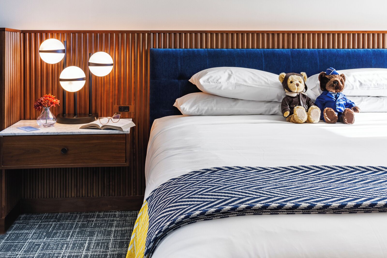 A hotel room with a wooden headboard, a bed with white linens, two teddy bears, a bedside table with a vase of flowers, and a patterned throw blanket.