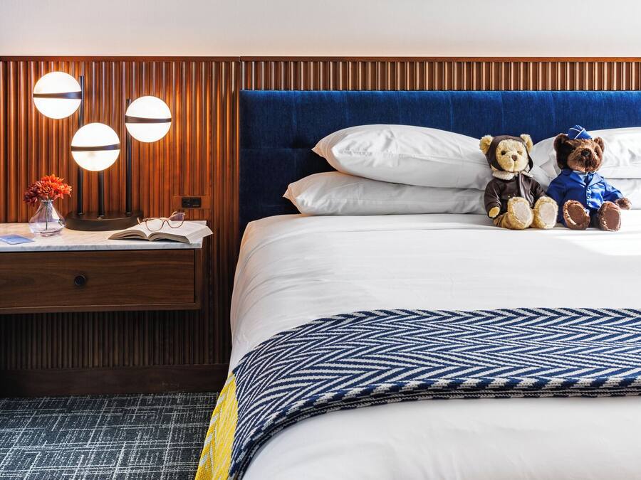 A hotel room with a wooden headboard, a bed with white linens, two teddy bears, a bedside table with a vase of flowers, and a patterned throw blanket.