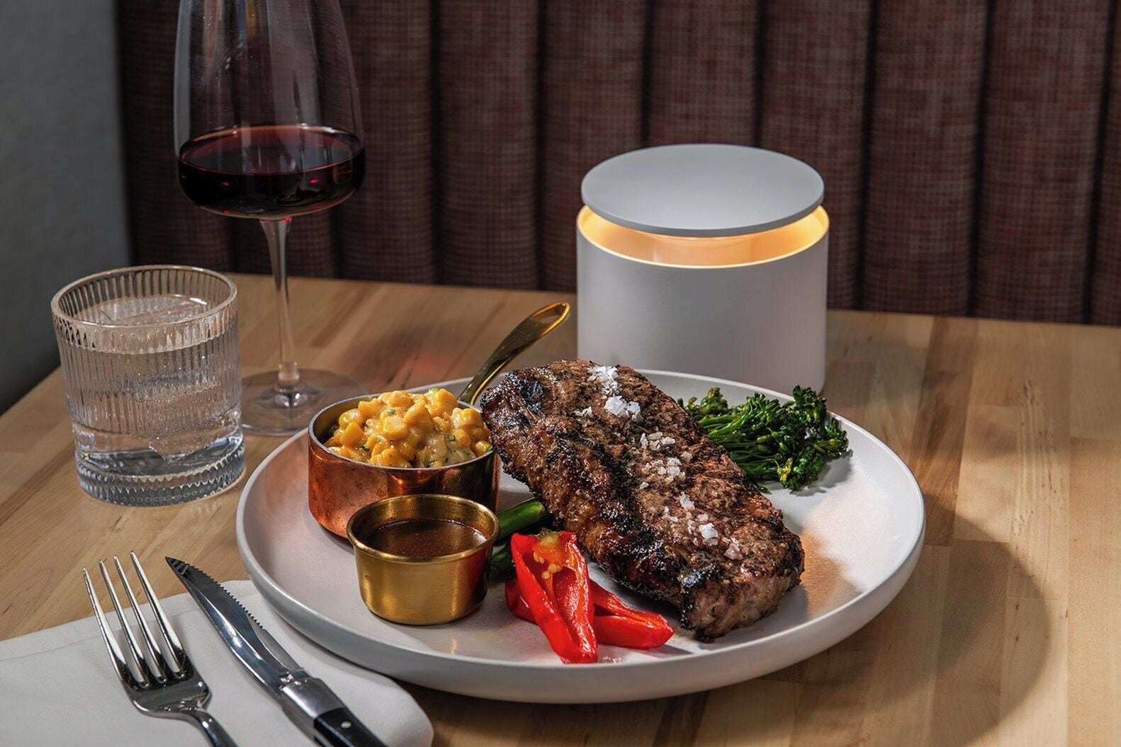 Une assiette contenant un steak, des légumes et un petit bol de macaronis au fromage, accompagnée d’un verre de vin rouge et d’une bougie posée sur une table.