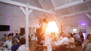 Une réception de mariage se déroule dans une grande salle aux hauts plafonds et aux poutres apparentes. Les invités sont assis à des tables rondes recouvertes de nappes blanches, et un couple danse au centre de la salle.